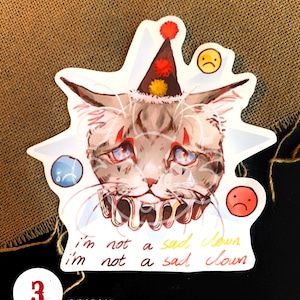 K&ouml;nnte beinhalten: Ein wei&szlig;er Aufkleber mit einer Comic-Katze, die einen roten und gelben Clownshut tr&auml;gt. Die Katze hat einen traurigen Ausdruck und weint. Der Text "i'm not a sad clown" steht unter der Katze. Der Aufkleber ist 7,5 cm x 7,3 cm gro&szlig;.