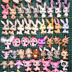 Peut inclure: Une collection de petites figurines d'animaux en plastique, principalement des lapins et des cochons, disposées en rangées sur un tapis de découpe vert avec des lignes de grille jaunes. Les figurines ont de grands yeux et diverses couleurs.