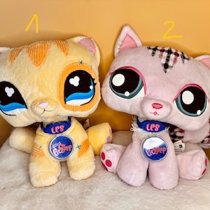 Könnte beinhalten: Zwei Plüschkatzenspielzeuge. Eines ist gelb mit orangefarbenen Streifen, das andere rosa mit einem Karomuster. Beide haben große, ausdrucksstarke Augen, blaue Halsbänder mit "LPS" und "Littlest Pet Shop"-Abzeichen.