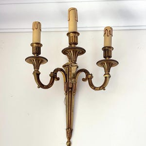 May include: A gold-toned wall sconce with three candle holders. The sconce has ornate detailing and curved arms. The candle holders have a classic design, and the candles are a cream color. The sconce is mounted on a white wall.