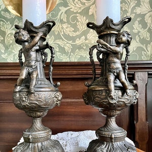 Könnte beinhalten: Ein Paar verzierte Kerzenleuchter aus Bronze mit Cherubfiguren, die die Kerzenhalter festhalten. Die Kerzenleuchter haben ein detailliertes Design mit aufwendigen Schnitzereien und einem Sockel mit einem gewellten Rand.