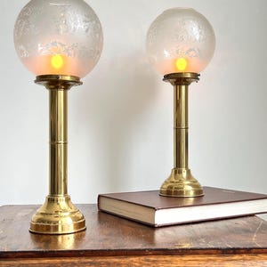 Op de afbeelding: Twee antieke messing tafellampen met matglazen bollen. De bollen hebben een delicaat bloemmotief en geven een warm, geel licht af. De lampen staan op een houten tafel, een ervan op een gesloten boek.