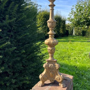 May include: A tall, ornate gold-colored candlestick holder. The base features decorative feet and floral accents. The candlestick has a detailed, layered design with a pointed top. The piece is outdoors, set against a backdrop of greenery and a blue sky.