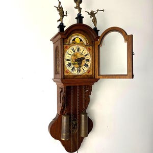 May include: A wooden wall clock with a decorative face, featuring Roman numerals and a detailed landscape scene. The clock has a pendulum and weights, and is adorned with three small bronze figures on top. The clock's open door reveals a mirrored interior.