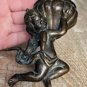 Op de afbeelding: Een bronzen cherubijnbeeldje dat een groot, rond vat vasthoudt. De cherubijn knielt met één vleugel uitgestrekt. Het beeldje heeft ingewikkelde details en een vintage esthetiek.