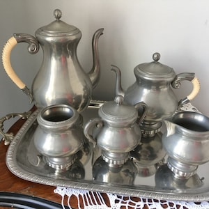 Peut inclure: Un service à thé en argent avec une théière, un pot à crème, un sucrier et un plateau. La théière a une poignée en bois et le service est sur une table en bois.