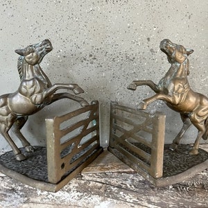 May include: A pair of antique brass bookends featuring rearing horses jumping over a fence. The bookends are in a distressed finish with a green patina.