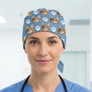 May include: A light blue scrub cap featuring a repeating pattern of brown tabby cat faces and white paw prints. The cap has a rounded crown and ties at the back, providing a secure fit for medical professionals in a clinical environment.