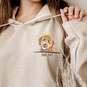 May include: A beige hooded sweatshirt featuring a graphic on the upper left chest. The graphic depicts a brown curly-haired dog with a yellow halo above its head, positioned above the text BRUNO in a black sans-serif font with a horizontal line.