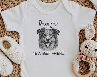 May include: A white short-sleeved baby bodysuit featuring a black ink-style illustration of a dog's face in the center, with the text Daisy's NEW BEST FRIEND printed above and below the dog. The bodysuit is accompanied by two white knit booties.