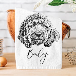 May include: A white kitchen towel featuring a black line art illustration of a curly-haired dog's face. Below the dog illustration, the name Bailey is printed in a black cursive script font. The towel is folded and displayed in a kitchen setting.