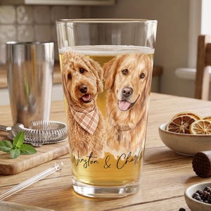 May include: A clear pint glass filled with a golden beverage, featuring a printed illustration of a curly-haired dog wearing a plaid bandana and a golden retriever, with the text Winston & Charlie written in black script below the dogs.