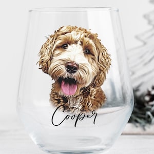 May include: A clear stemless wine glass featuring a detailed illustration of a brown and white curly-haired dog with its tongue out. The name Cooper is printed in black cursive script lettering directly below the dog's portrait on the glass.