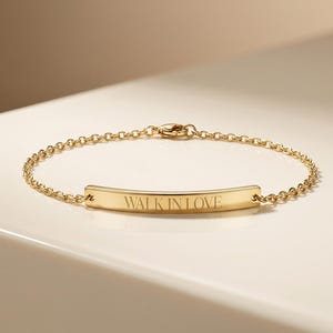May include: A gold bracelet featuring a fine chain and a curved rectangular bar. The bar is inscribed with the words "WALK IN LOVE". The bracelet is presented on a white surface.