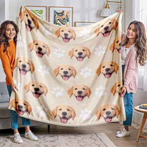 Op de afbeelding: Een beige plaid met een herhalend patroon van golden retriever-gezichten met open bekken en uitgestoken tongen, afgewisseld met witte pootafdrukken. De deken wordt omhoog gehouden om de volledige grafische print te tonen.
