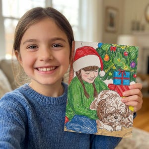 May include: A young girl smiles, holding a colorful Christmas-themed drawing. The drawing depicts a girl in a Santa hat, a dog, a decorated Christmas tree, and wrapped gifts. The girl in the image is wearing a blue sweater.