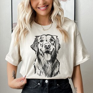Beige t-shirt featuring a custom black-and-white sketch of a happy dog. Worn by a smiling woman paired with black jeans for a casual look. A perfect personalized gift for dog lovers or pet parents, combining style and a unique design.