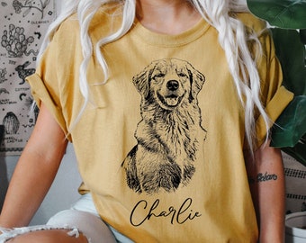 May include: A mustard yellow short-sleeve t-shirt featuring a black line-art illustration of a dog sitting and looking forward. Below the dog illustration, the name Charlie is printed in a black cursive script font on the front of the shirt.