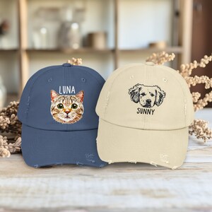 May include: A navy blue baseball cap featuring a colorful cat illustration and the text LUNA, alongside a beige baseball cap featuring a black and white dog illustration and the text SUNNY. Both caps have a distressed, frayed brim design.