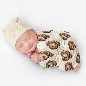 May include: A swaddled infant wearing a cream-coloured knit beanie with a cable pattern. The swaddle blanket is light beige and features a repeating pattern of brown puppy faces and white paw prints across the entire fabric surface.