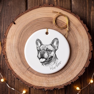 May include: A round white ceramic ornament featuring a black and white sketch of a French Bulldog with its tongue out. The name Winston is written in cursive script below the dog. A gold string is attached to the top for hanging.