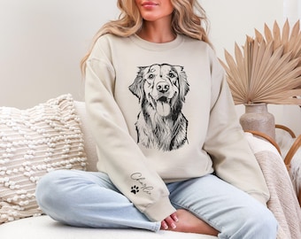 May include: A beige crewneck sweatshirt featuring a black line art illustration of a dog on the chest. The left sleeve includes the name Charlie in black cursive script above a small black paw print icon, all printed on a light fabric.