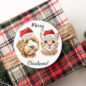 May include: A round Christmas gift tag featuring a dog and a cat wearing Santa hats. The dog's name is Cooper, and the cat's name is Luna. The tag is on a plaid gift box wrapped with twine, with the words "Merry Christmas!"
