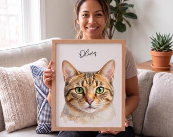 May include: A framed portrait print featuring a watercolor-style illustration of a brown tabby cat with green eyes. The name Oliver is printed in black script above the cat's head. The artwork is encased in a light-colored wooden frame.