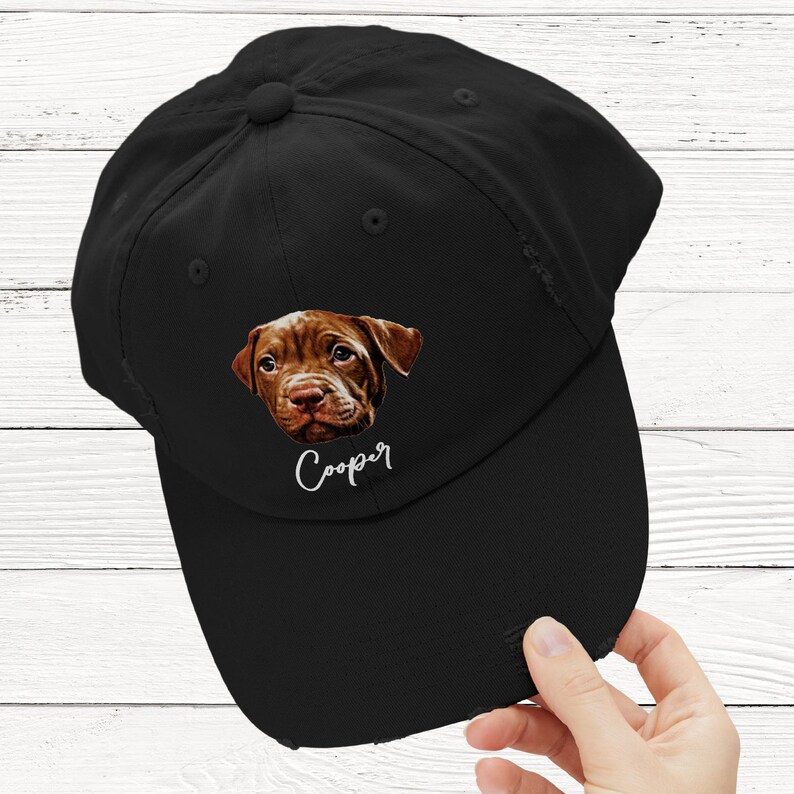 May include: A black baseball cap with a distressed brim and a graphic of a brown puppy face centered on the front. Below the puppy image, the name Cooper is printed in a white cursive script font. The cap features a button on the top crown.