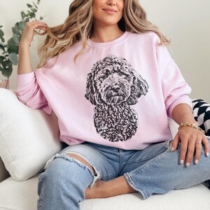 K&ouml;nnte beinhalten: Eine Frau tr&auml;gt ein rosa Sweatshirt mit einer schwarz-wei&szlig;en Illustration eines Hundes auf der Vorderseite. Sie sitzt auf einem wei&szlig;en Sofa mit einer schwarz-wei&szlig;en karierten Wolldecke.