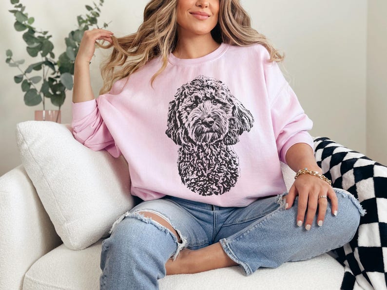 K&ouml;nnte beinhalten: Eine Frau tr&auml;gt ein rosa Sweatshirt mit einer schwarz-wei&szlig;en Illustration eines Hundes auf der Vorderseite. Sie sitzt auf einem wei&szlig;en Sofa mit einer schwarz-wei&szlig;en karierten Wolldecke.