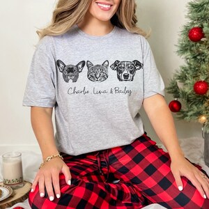 May include: A gray t-shirt with a black and white illustration of three pet faces. The text "Charlie, Luna & Bailey" is printed below the illustrations. The person wearing the shirt is sitting on a red and black plaid blanket.