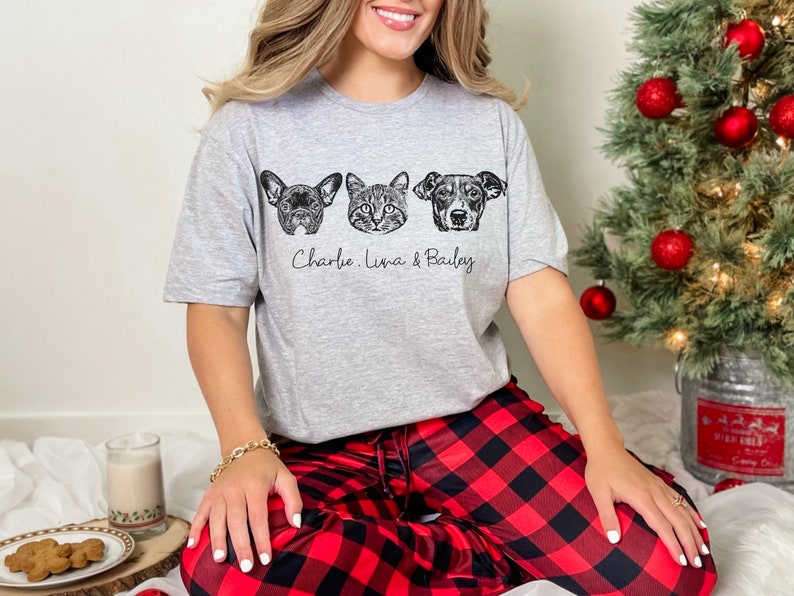 May include: A gray t-shirt with a black and white illustration of three pet faces. The text "Charlie, Luna & Bailey" is printed below the illustrations. The person wearing the shirt is sitting on a red and black plaid blanket.