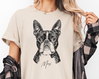 May include: A beige short-sleeved t-shirt featuring a black and white graphic illustration of a Boston Terrier dog's face. Below the dog illustration, the name Max is printed in a black cursive script font. The shirt has a crew neckline.