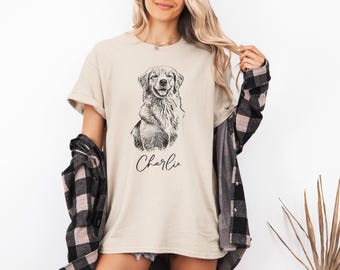 May include: A beige short-sleeved t-shirt featuring a black line-art illustration of a dog on the chest, with the name Charlie written in a cursive script font below the dog graphic, worn with a black and beige plaid long-sleeved button-down shirt.