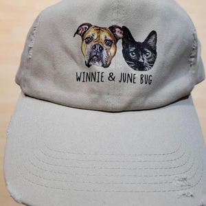 May include: A light beige baseball cap with a distressed brim and a graphic print featuring a brown dog and a black cat. Below the animal portraits, the text "WINNIE & JUNE BUG" is printed in a simple black font on the front panel of the cap.