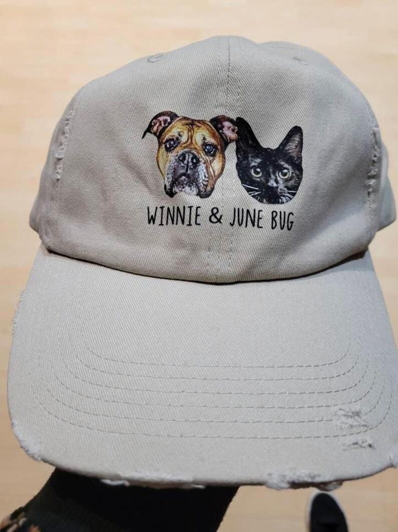 May include: A light beige baseball cap with a distressed brim and a graphic print featuring a brown dog and a black cat. Below the animal portraits, the text "WINNIE & JUNE BUG" is printed in a simple black font on the front panel of the cap.