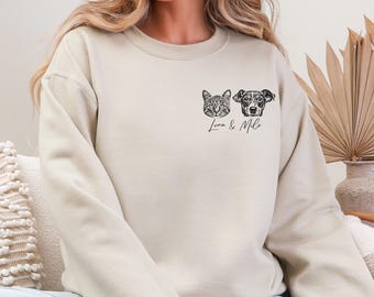 May include: A beige crewneck sweatshirt featuring a black line art graphic on the left chest. The design depicts a cat and a dog face side-by-side with the handwritten-style text "Luna & Milo" printed directly underneath the animal portraits.
