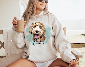 May include: A beige hooded sweatshirt featuring a graphic of a brown and white doodle dog with its tongue out, set against a light blue watercolor splash. The name Cooper is written in black cursive script below the dog illustration.