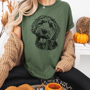 May include: A woman wearing a green t-shirt with a black and white illustration of a doodle dog. She is wearing a brown cardigan and black pants.