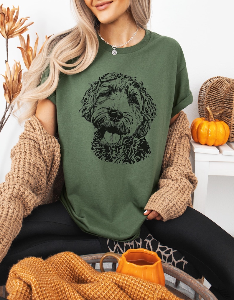 May include: A woman wearing a green t-shirt with a black and white illustration of a doodle dog. She is wearing a brown cardigan and black pants.