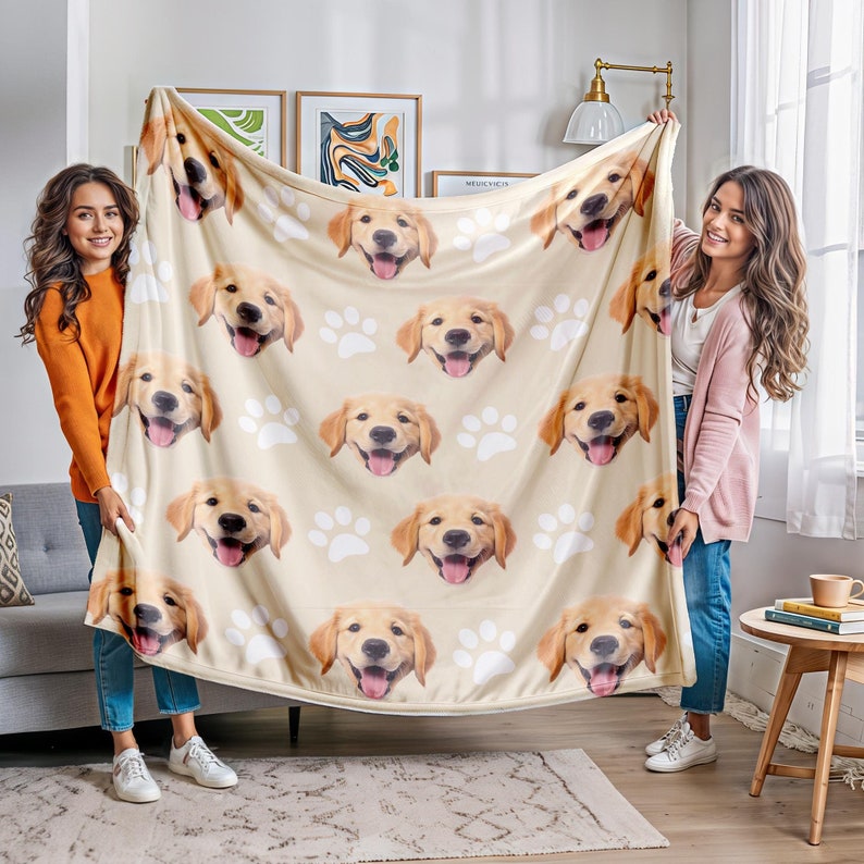 Op de afbeelding: Een beige plaid met een herhalend patroon van golden retriever-gezichten met open bekken en uitgestoken tongen, afgewisseld met witte pootafdrukken. De deken wordt omhoog gehouden om de volledige grafische print te tonen.
