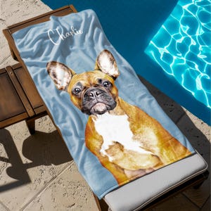 May include: A light blue beach towel featuring a detailed image of a French Bulldog. The dog has brown and white fur and is lying on a wooden lounge chair. The name "Charlie" is written in white script on the towel.