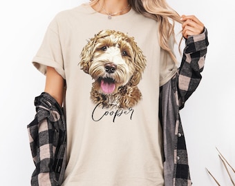 May include: A beige short-sleeved t-shirt featuring a detailed illustration of a brown and white doodle dog with its tongue out. Below the dog, the name Cooper is printed in a black cursive script. A black and beige plaid flannel shirt is worn over it.