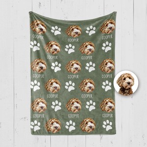 Op de afbeelding: Een saliegroene fleece deken met een herhalend patroon van een bruin-witte doodle hondenkop, witte pootafdrukken en de naam COOPER in witte letters. Een cirkelvormige inzet toont hetzelfde hondengezicht op een witte achtergrond.