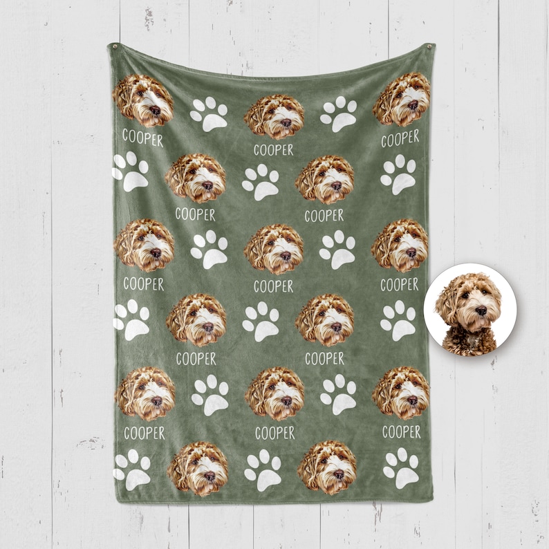 Op de afbeelding: Een saliegroene fleece deken met een herhalend patroon van een bruin-witte doodle hondenkop, witte pootafdrukken en de naam COOPER in witte letters. Een cirkelvormige inzet toont hetzelfde hondengezicht op een witte achtergrond.