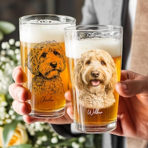 May include: Two clear glass pint glasses filled with golden beer. The left glass features a black line-art illustration of a curly-haired dog with the name Charlie below. The right glass displays a realistic portrait of a dog with the name Willow.