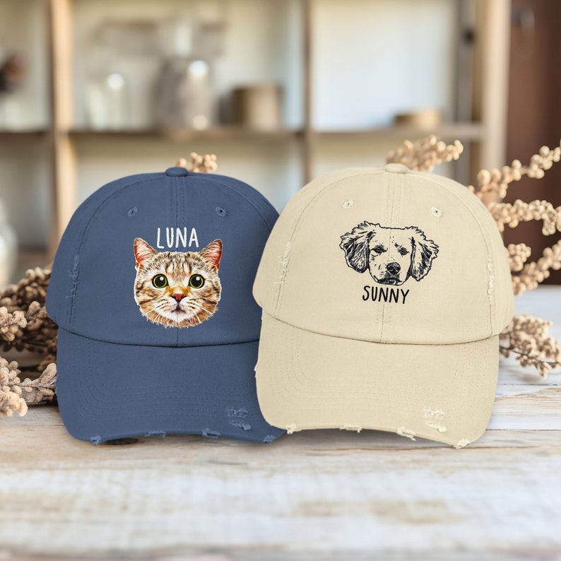 May include: A navy blue baseball cap featuring a colorful cat illustration and the text LUNA, alongside a beige baseball cap featuring a black and white dog illustration and the text SUNNY. Both caps have a distressed, frayed brim design.