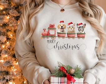 May include: A cream-colored crewneck sweatshirt featuring a graphic of a red train with three carriages. Each carriage holds a dog or cat wearing a red Santa hat. Below the train, the text reads Merry Christmas in black cursive and print font.
