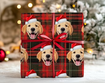 May include: A square gift box wrapped in red and black plaid paper featuring a repeating pattern of golden retriever dog faces wearing red and white Santa hats. The package is tied with a thin, smooth red ribbon centered in a bow on the front.